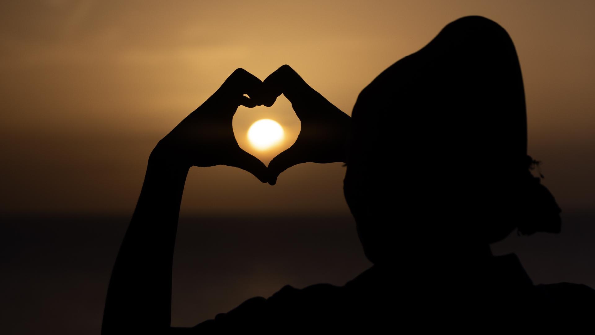 A person forming a heart with their hands around the sunset.
