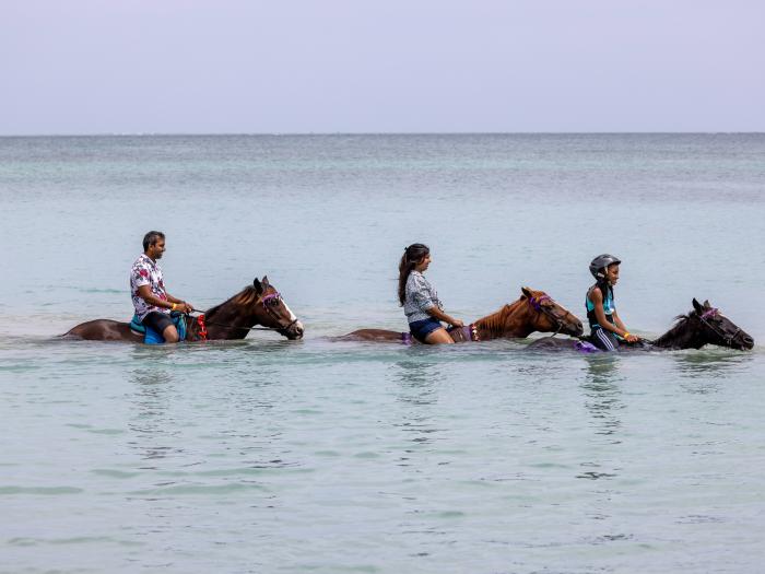 Three persons wading through Buccoo Beach on horseback